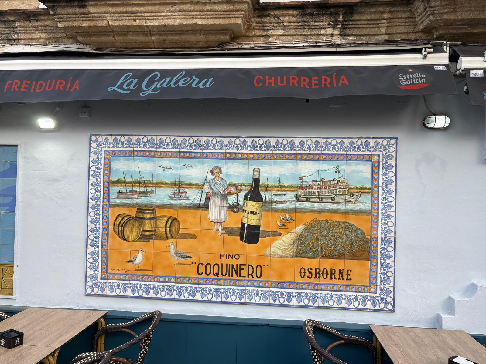 Azulejo pintado a mano del Fino Coquinero de Osborne en la fachada de La Galera: mujer con bandeja de pescado, barcas y redes junto al Guadalete.